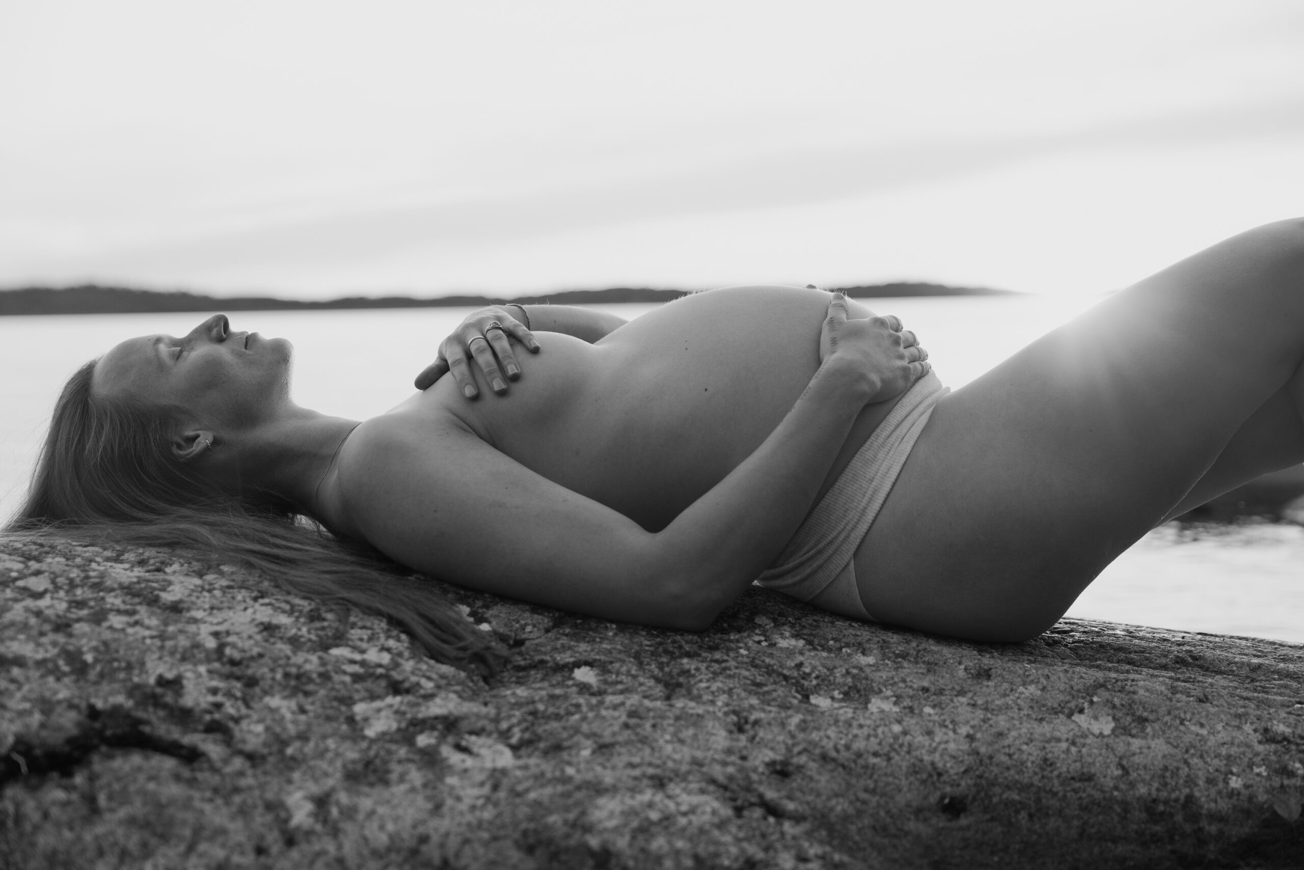 Svartvit bild på gravid kvinna som ligger på klipporna vid havet.