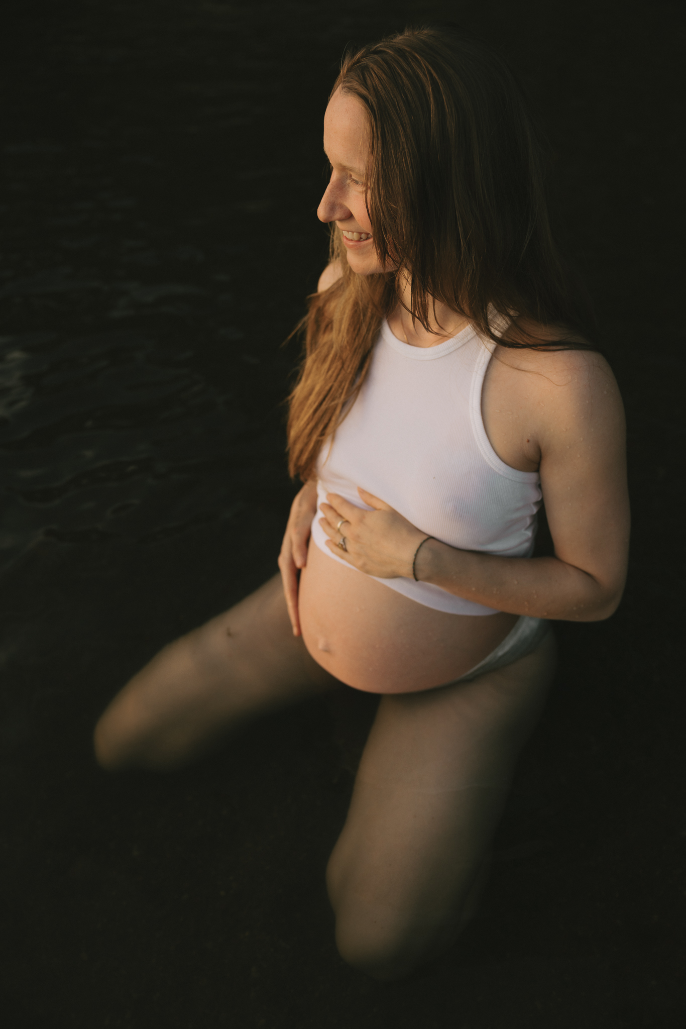 En gravid kvinna iklädd linne och trosor sitter i vattnet och håller händerna på magen.